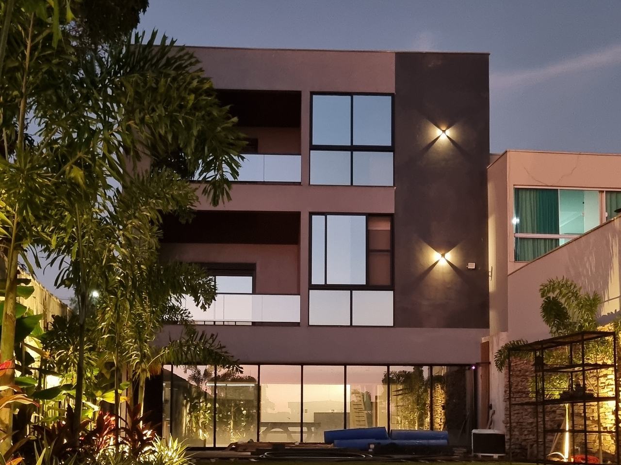 Residência Deck 519 ao entardecer: três pavimentos com fachada escura, panos de vidro e jardim iluminado